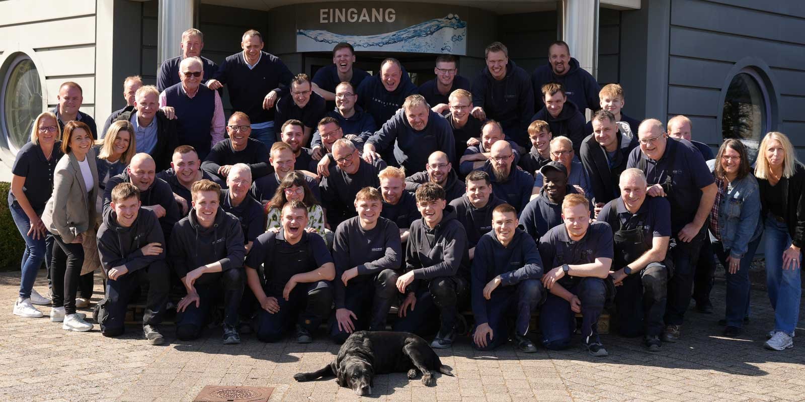 Eine große Gruppe von Menschen steht fröhlich vor einem Gebäude, während ein schwarzer Hund entspannt im Vordergrund liegt.