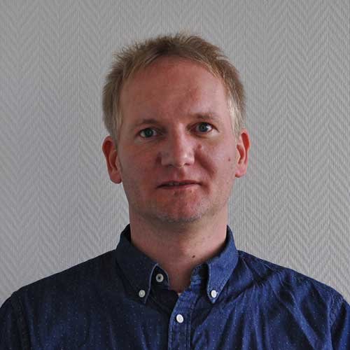 Ein Mann mit blonden Haaren und einem blauen Hemd steht vor einer hellen, strukturierten Wand und schaut direkt in die Kamera.