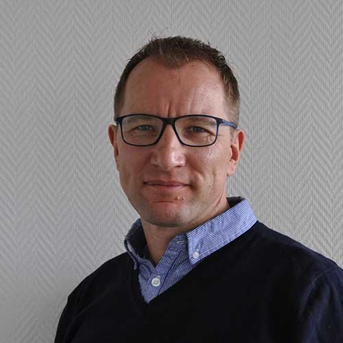 Ein Mann mit Brille und einem dunklen Pullover steht vor einer hellen, strukturierten Wand und schaut direkt in die Kamera.