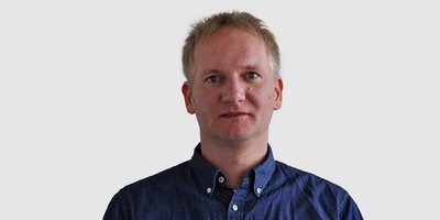 Ein Mann mit blonden Haaren und einem blauen Hemd steht vor einem neutralen Hintergrund und schaut direkt in die Kamera.