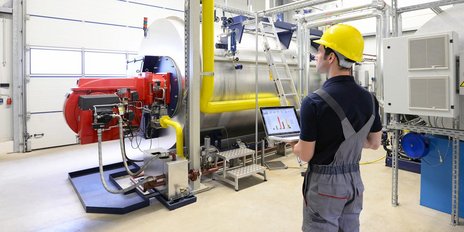Ein Mann mit Bauhelm steht mit einem Laptop vor einer Anlage